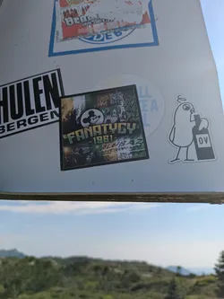 Street sticker The central sticker is a multi-layered design. Its upper left section features a dark, somewhat menacing Mickey Mouse-like figure set against a background that resembles the Irish flag (green, white, and orange vertical stripes). To its right, a detailed illustration of a historical European building, possibly a castle or cathedral, is depicted against a golden or yellowish background. The central text "FANATYCY" is prominently displayed in green with a white outline, followed by "1981-" and what appears to be "ALUBAZ" below it. The lower part of the sticker shows a dynamic scene of a large crowd with raised arms, possibly supporters at a stadium or a protest, with flares or smoke adding to the intense atmosphere. The overall aesthetic suggests a connection to football fan culture, specifically ultra groups.