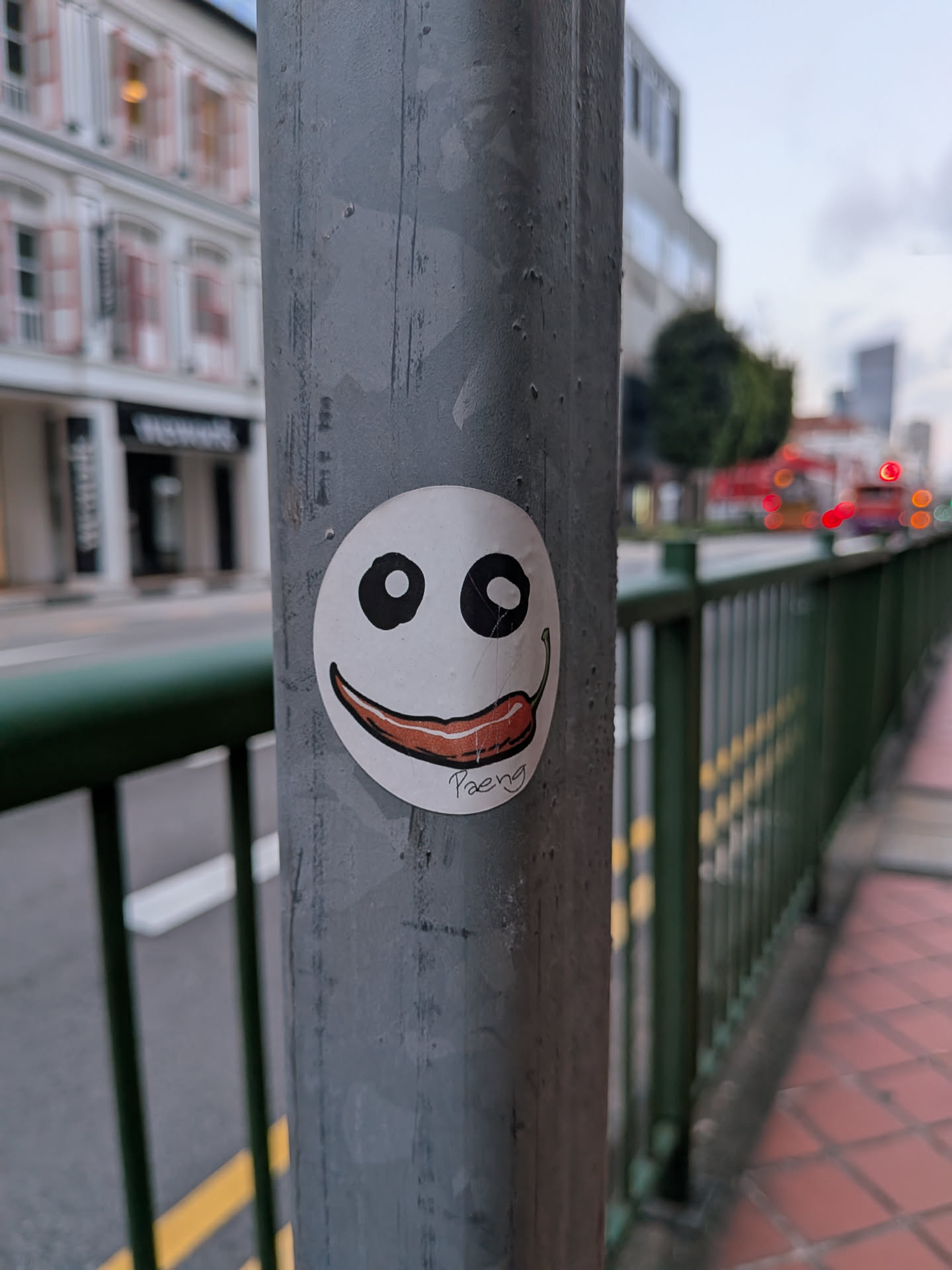 A round white sticker featuring a simple smiley face. The eyes are large black circles with white dots, and the mouth is depicted as a red chili pepper with a green stem. The name 'Paeng' is written in black script at the bottom.