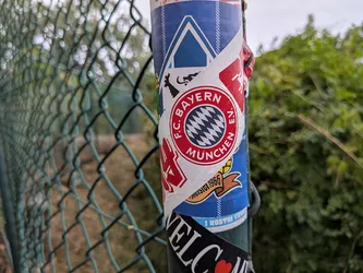 Street sticker A circular red and white FC Bayern München logo sticker layered over a blue plaid sticker on a green metal pole next to a chain-link fence.