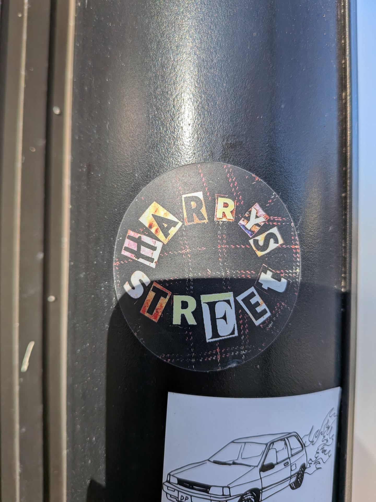 The sticker is circular and features a plaid background with red and white lines. The text is arranged in a circular pattern, with letters cut out from various patterned papers, including some with floral and abstract designs. The letters are white with black outlines and appear to be from magazine clippings. The text reads 'HARRY'S STREET' in a fragmented, collage-like style.