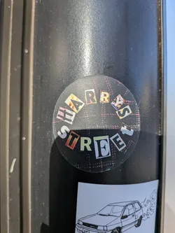 Street sticker The sticker is circular and features a plaid background with red and white lines. The text is arranged in a circular pattern, with letters cut out from various patterned papers, including some with floral and abstract designs. The letters are white with black outlines and appear to be from magazine clippings. The text reads 'HARRY'S STREET' in a fragmented, collage-like style.