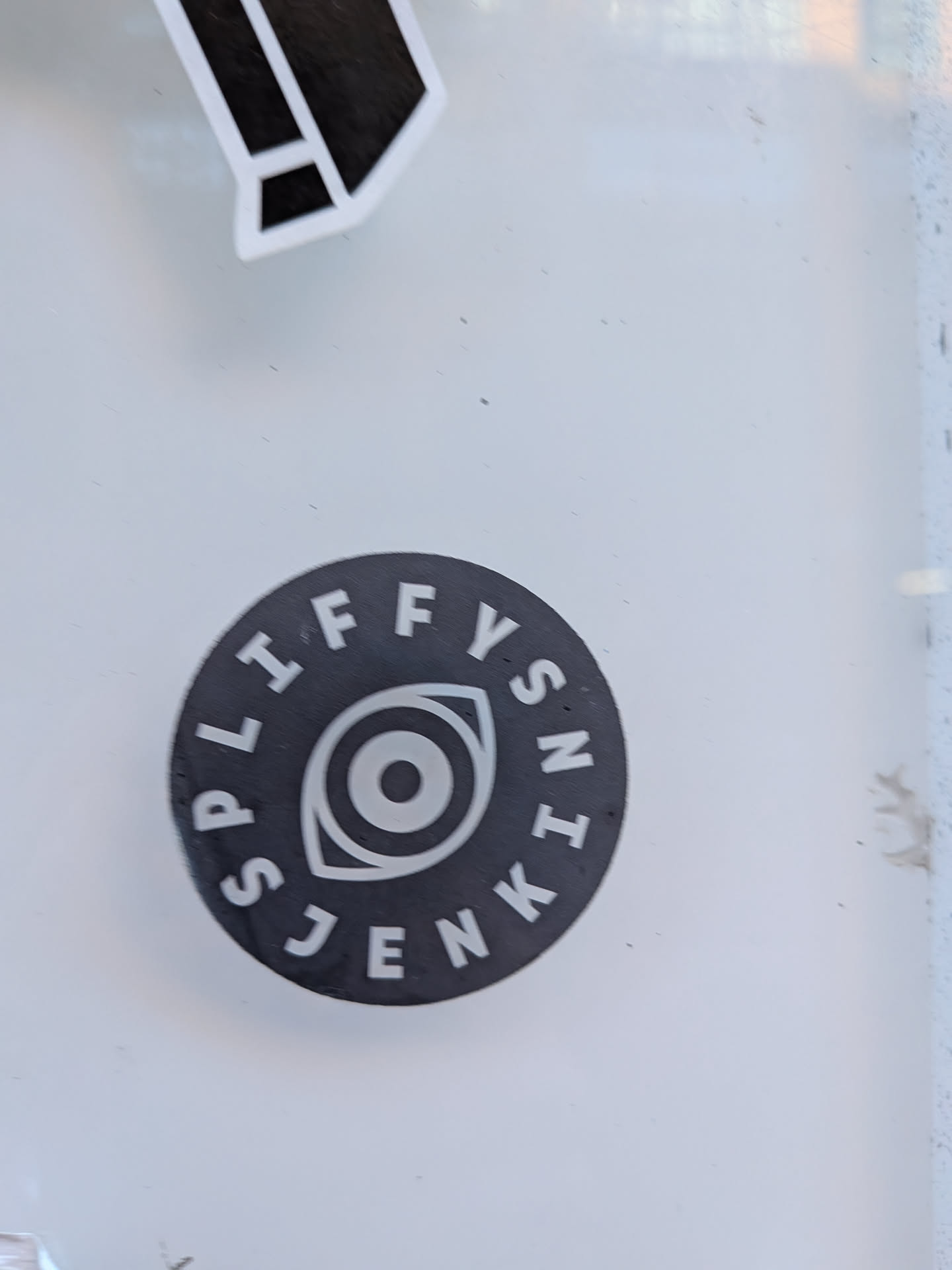 A circular sticker with a dark grey or black background. In the center, there is a stylized eye symbol rendered in white, featuring an almond shape, an inner circle for the iris, and a central dot for the pupil. Around the perimeter of the circle, white uppercase sans-serif letters spell out "SPIFFY SNK JENK", following the curve of the sticker's edge. The overall design is clean, high-contrast, and minimalist.