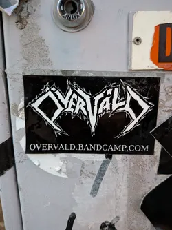 Street sticker The sticker is rectangular with a black background. At the top, it features a stylized band logo in white, with sharp, angular lettering that resembles gothic or heavy metal typography. Below the logo, in smaller white text, is the URL 'OVERVALD.BANDCAMP.COM'. The sticker appears to be slightly worn and has a glossy finish.