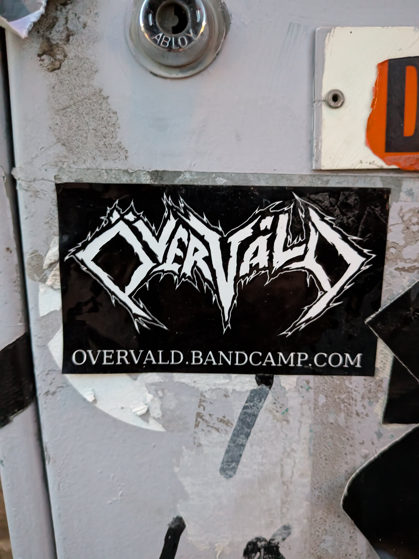 The sticker is rectangular with a black background. At the top, it features a stylized band logo in white, with sharp, angular lettering that resembles gothic or heavy metal typography. Below the logo, in smaller white text, is the URL 'OVERVALD.BANDCAMP.COM'. The sticker appears to be slightly worn and has a glossy finish.