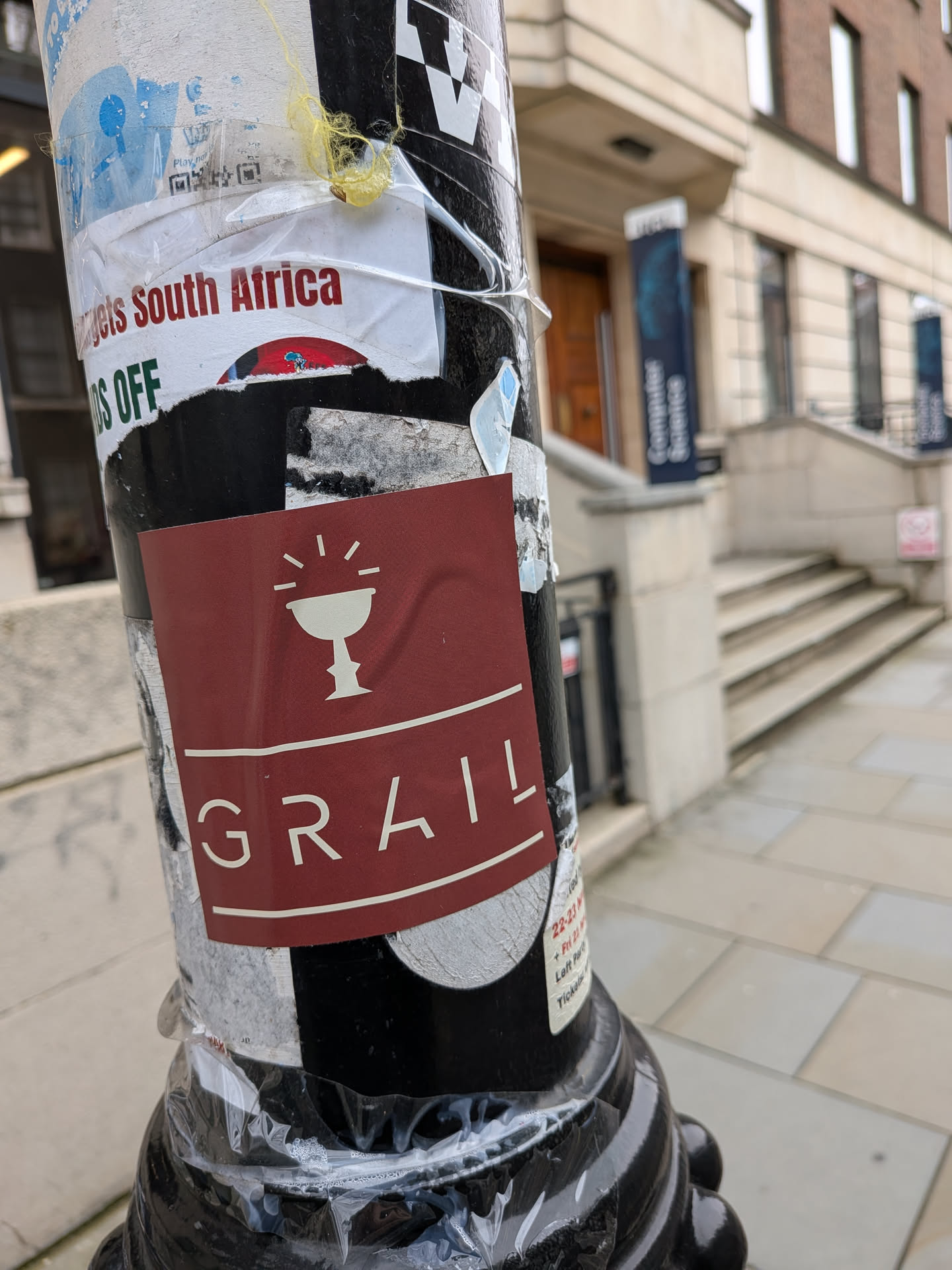A rectangular sticker with rounded corners, featuring a deep maroon background. At the top, a white outline illustration of a chalice or goblet is depicted, with short radiating lines above it, suggesting importance or light. Below the chalice, a thin white horizontal line separates it from the word "GRAIL," which is written in large, uppercase, sans-serif white letters. Another thin white horizontal line is present below the text. The sticker appears slightly creased, conforming to the curved surface of the pole.