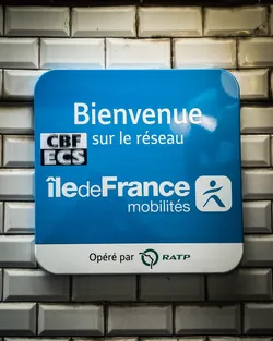 Street sticker A rectangular sign with rounded corners, featuring a bright blue upper section and a white lower section. The top blue part displays 'Bienvenue sur le réseau' in large white sans-serif font. Below this, a small black rectangular box contains 'CBF ECS' in white, horizontally striped text. Further down, 'Île-de-France mobilités' is written in white, with a stylized 'i' in 'Île'. To the right of 'mobilités' is a white square icon depicting a dark blue/black stylized human figure in motion. The bottom white section of the sign reads 'Opéré par' in dark grey text, followed by the green RATP logo, which is a stylized profile of a face within a 'Q' shape, and the word 'RATP' in green. The sign is affixed to a wall of glossy, rectangular white subway tiles with dark grout lines.