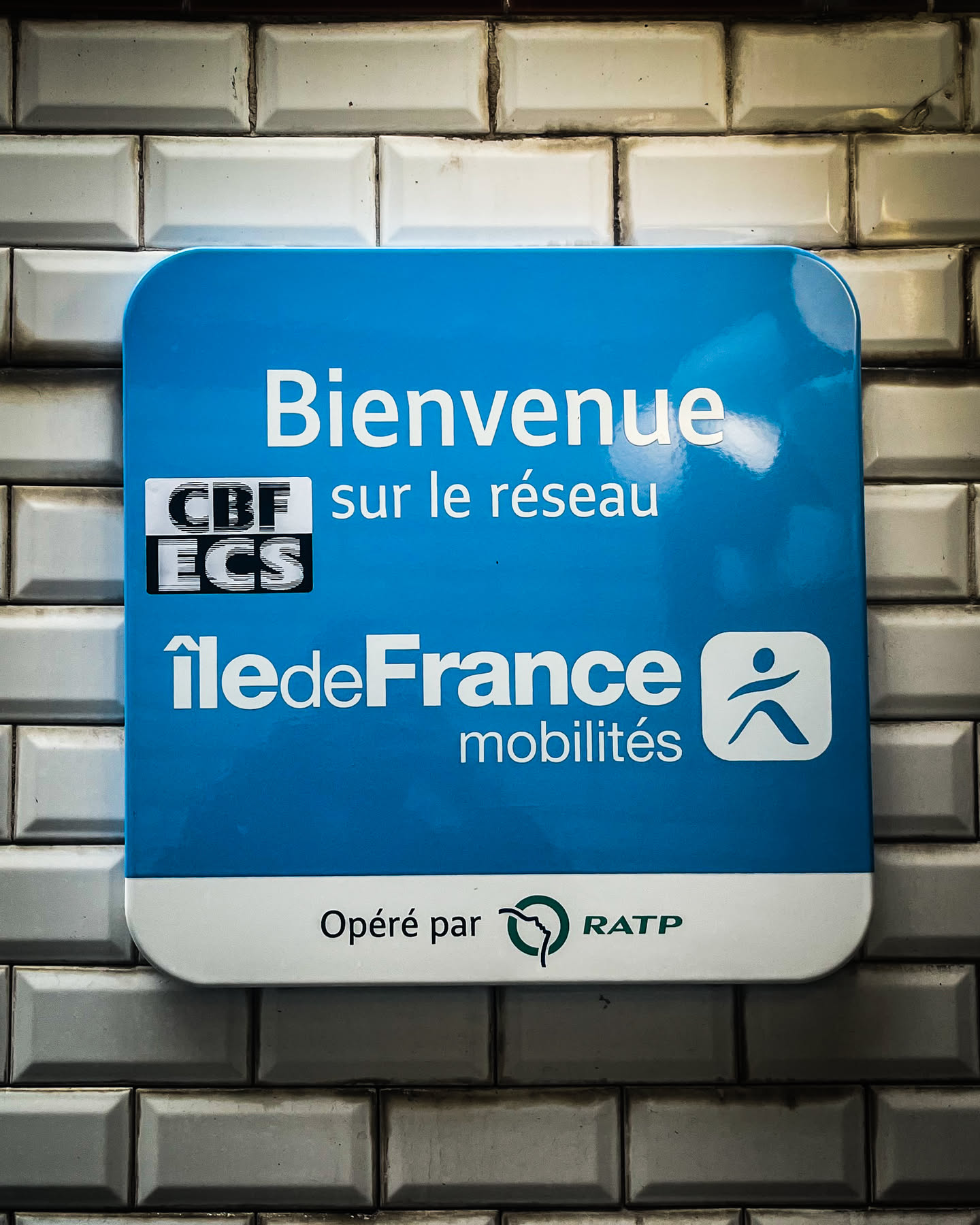 A rectangular sign with rounded corners, featuring a bright blue upper section and a white lower section. The top blue part displays 'Bienvenue sur le réseau' in large white sans-serif font. Below this, a small black rectangular box contains 'CBF ECS' in white, horizontally striped text. Further down, 'Île-de-France mobilités' is written in white, with a stylized 'i' in 'Île'. To the right of 'mobilités' is a white square icon depicting a dark blue/black stylized human figure in motion. The bottom white section of the sign reads 'Opéré par' in dark grey text, followed by the green RATP logo, which is a stylized profile of a face within a 'Q' shape, and the word 'RATP' in green. The sign is affixed to a wall of glossy, rectangular white subway tiles with dark grout lines.