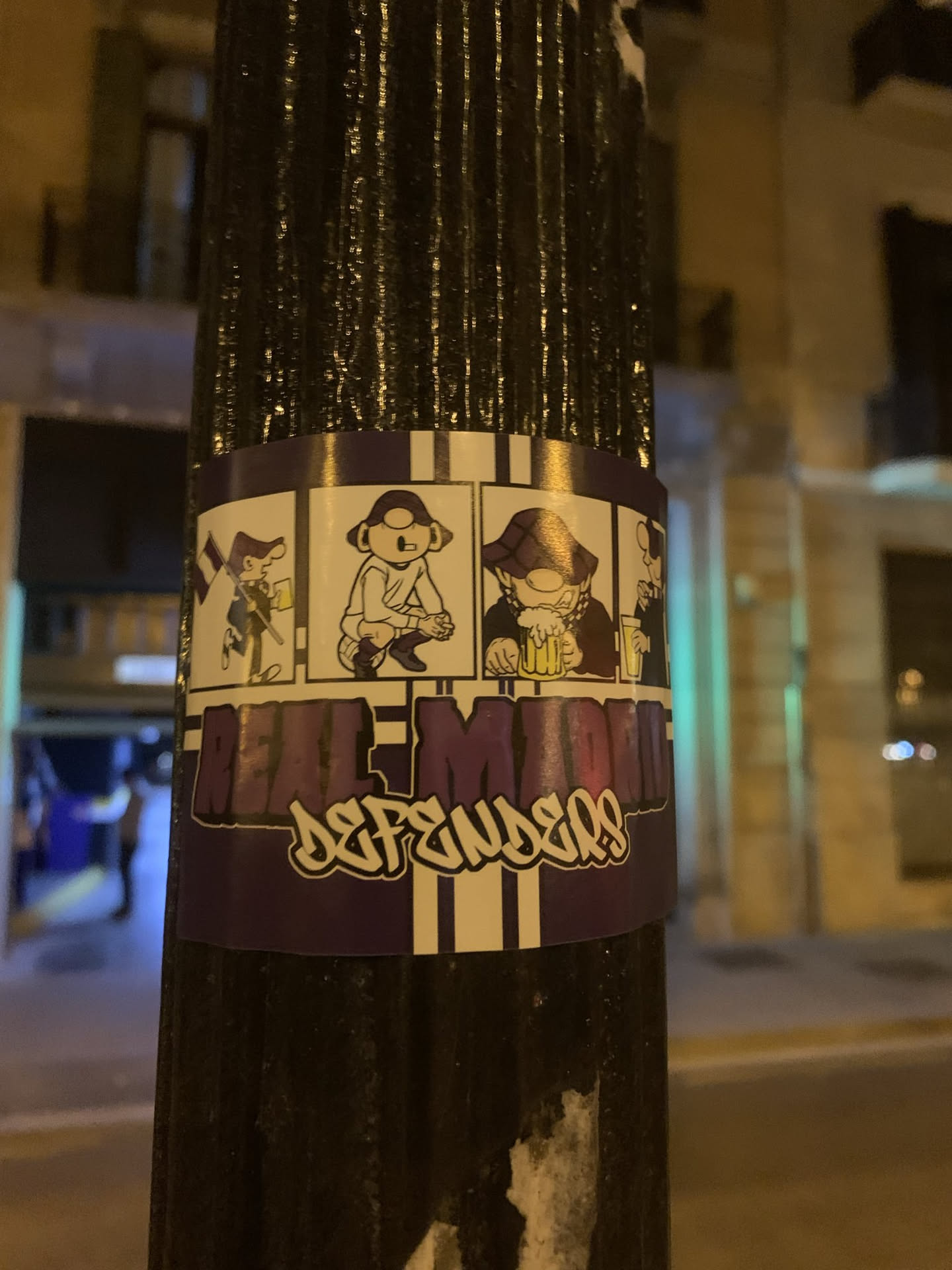 The sticker features a vibrant purple and white striped background, with a central white panel showcasing four distinct cartoon characters. From left to right, the first character holds a striped flag and a beer, the second plays an electric guitar, the third (wearing a hat) enthusiastically drinks from a large foamy beer mug, and the fourth (partially visible) also appears to be enjoying a drink. Below the character panels, the words 'REAL MADRID' are prominently displayed in large, purple, blocky letters, partially overlaid by the word 'DEFENDERS' in a white, graffiti-style font with a black outline.