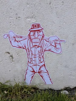 Street sticker A white sticker with red line art depicting a stylized male character. The figure has a large, wide grin showing many teeth, a prominent nose, and defined eyes. He wears a hat with the letters 'SKB' printed on it, a collared shirt with suspenders, and pants. He is holding a stick or club horizontally across his shoulders with both hands. The sticker is placed on a textured, off-white concrete wall, with green grass visible at the bottom.