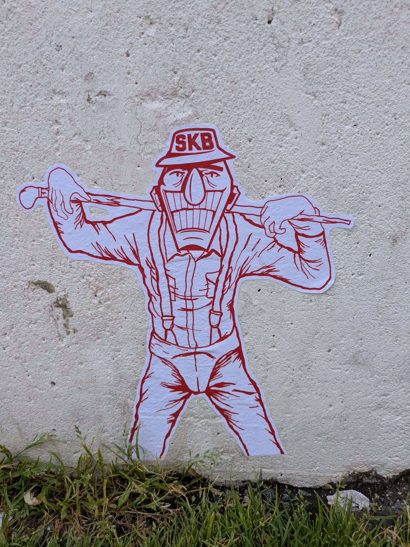 A white sticker with red line art depicting a stylized male character. The figure has a large, wide grin showing many teeth, a prominent nose, and defined eyes. He wears a hat with the letters 'SKB' printed on it, a collared shirt with suspenders, and pants. He is holding a stick or club horizontally across his shoulders with both hands. The sticker is placed on a textured, off-white concrete wall, with green grass visible at the bottom.