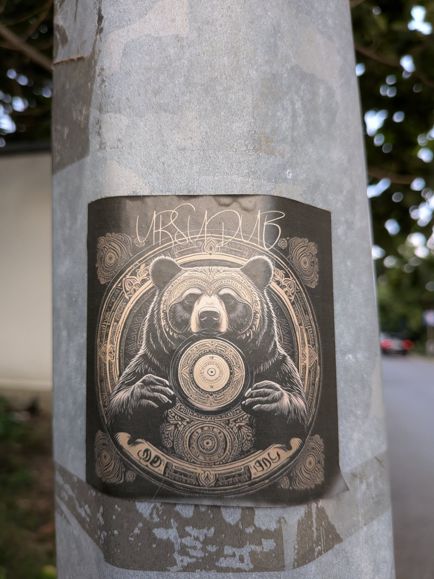 A black and gold square sticker featuring a detailed illustration of a bear holding a vinyl record. The bear is centered within an intricate, circular mandala frame. Stylized text 'URSUDUB' is written at the top, and a banner at the bottom contains additional markings.