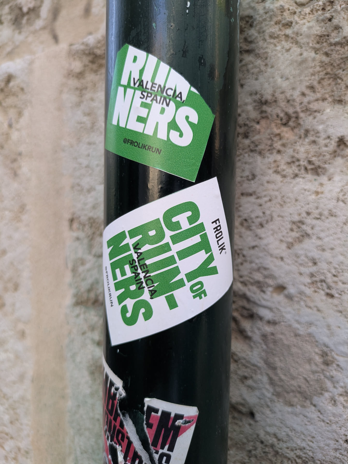 This image shows a pole with several stickers on it. The main sticker in focus is green and white. It says "Valencia Runners" with Valencia, Spain as its location. The sticker is also labeled with "@Frolikrun". There is a second similar sticker that says "City of Valencia Runners" and also labeled with Frolikrun. The pole appears to be made of metal and is mounted on a stone textured background.