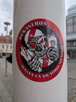 Street sticker The sticker is circular with a red border. At its center is a detailed illustration of a skull wearing a helmet with a red and white striped pattern. The skull has glowing red eyes and a menacing grin. It is holding a bloody knife in its left hand and a lit bomb in its right hand. The background within the circle is black, with some white accents. Around the top of the circle, in white text with a black outline, are the words 'BUKANEROS'. Along the bottom of the circle, also in white text with a black outline, are the words 'HASTA LA VICTORIA SIEMPRE'. There is a small crest-like emblem on the skull's chest.