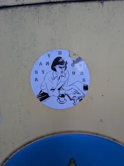 Street sticker The sticker is circular and features a black and white illustration of a woman sitting at a desk, looking intently at a book. She is wearing a collared shirt and has short, dark hair. To her left, there is a coffee cup and saucer, and to her right, a small glass. The background of the sticker is a light blue. Around the edge of the circle, the letters 'T H E A N S T A G O U R S' are arranged in a circular pattern. There is a small signature at the bottom left of the illustration that appears to read 'Kajsa Damelien'. The sticker shows some signs of wear and tear, with a small piece missing from the top edge.