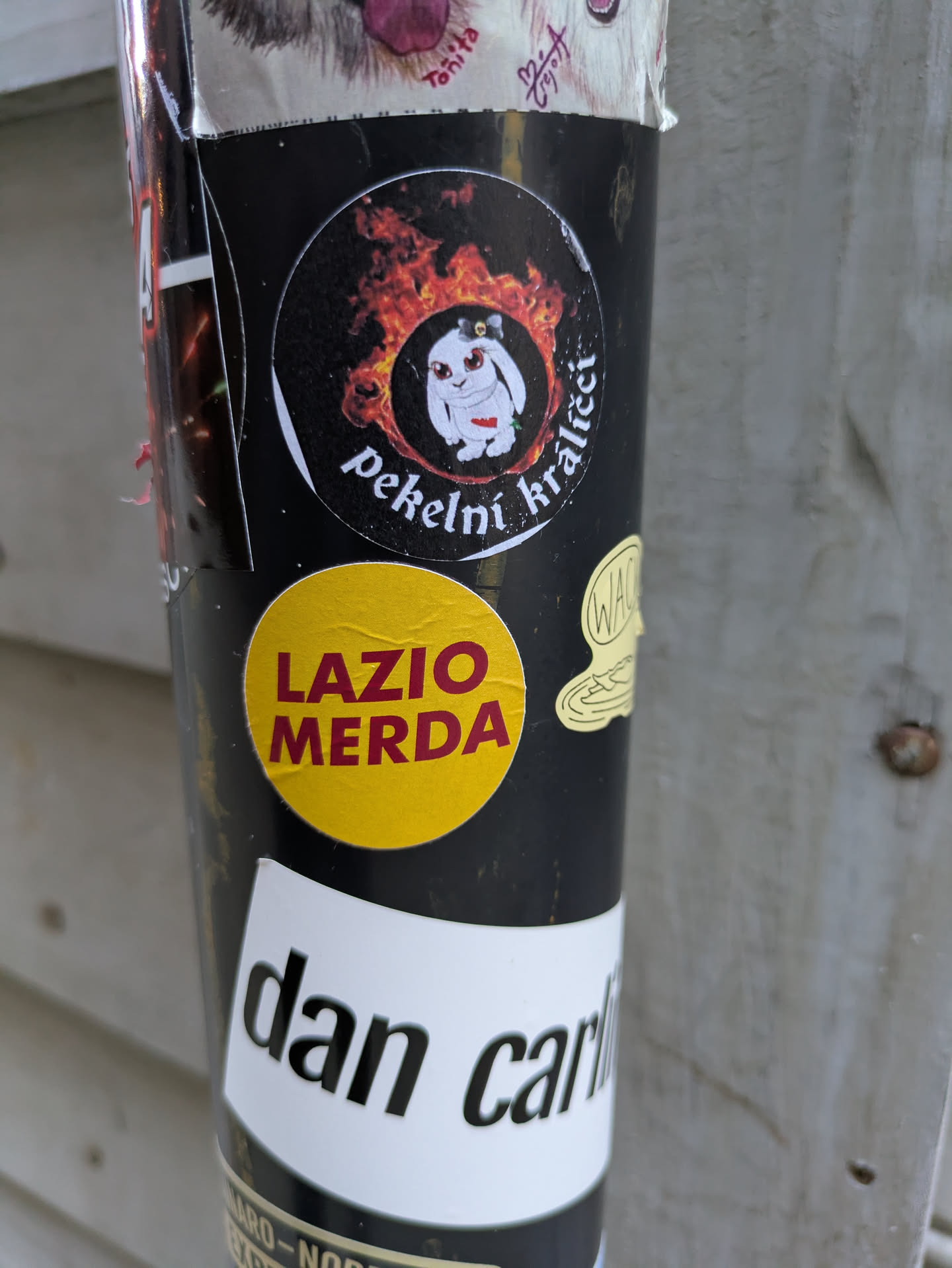 The central focal point is a black circular sticker featuring a white cartoon creature, resembling a rabbit or bunny, with prominent red eyes and a small red heart on its chest. This creature is depicted wearing a small grey hat and is engulfed in vibrant orange and red flames. Below this, there is a bright yellow circular sticker with the bold red text "LAZIO MERDA." Further down, a white rectangular sticker is partially visible with the black text "dan carl" cut off.