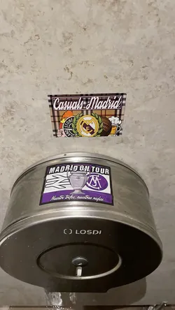 Street sticker The sticker features the text 'Casuals Madrid' in a stylized white font with a purple outline, set against a background resembling a brown and black plaid pattern. Below the text, from left to right, are several distinct elements: a hand holding a torch with the 'STONE ISLAND' compass logo, the circular crest of Real Madrid in yellow and purple, a brown vintage-style football, and a frothy mug of beer. The overall design combines elements of football fan culture, fashion, and leisure.