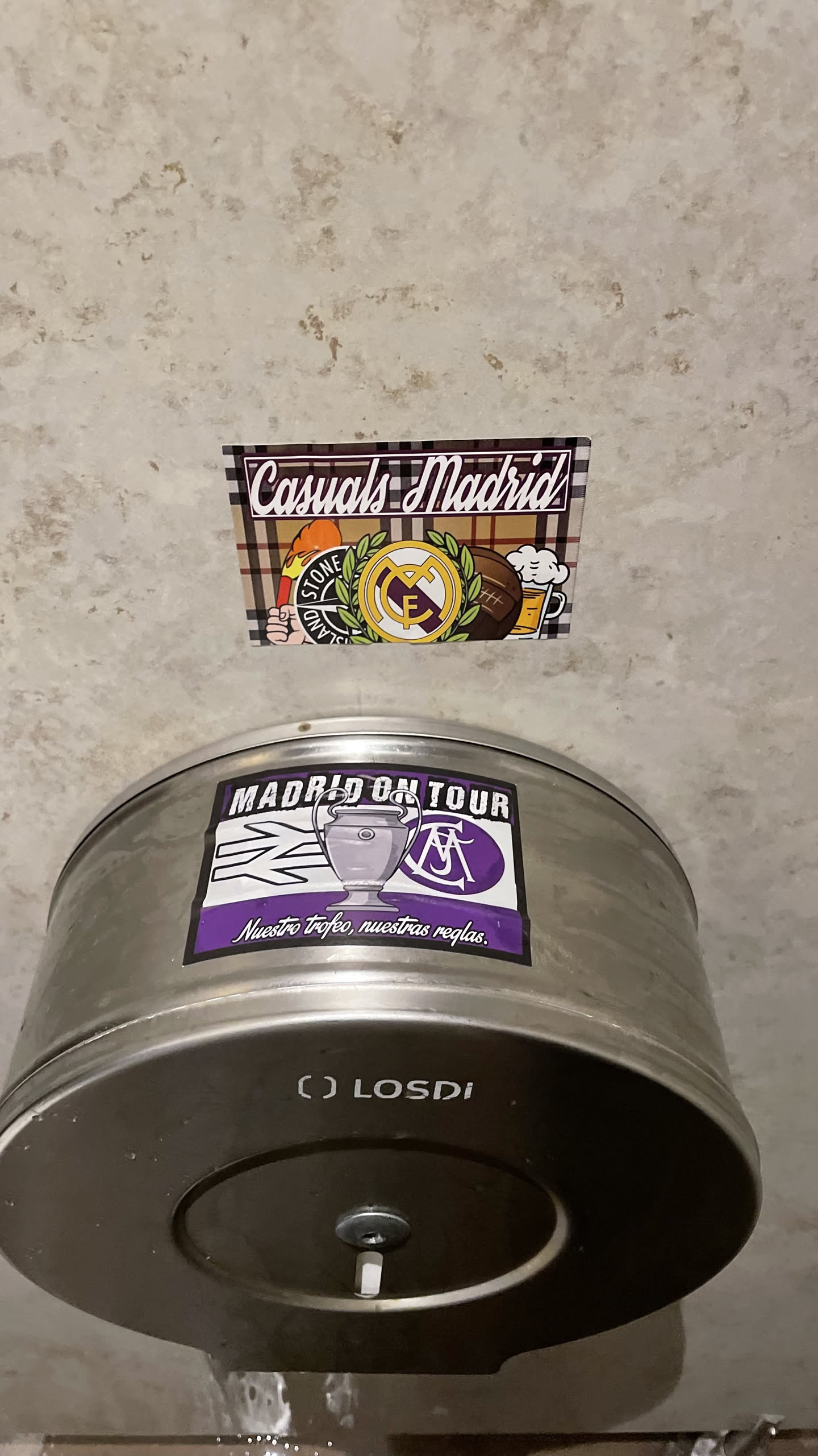 The sticker features the text 'Casuals Madrid' in a stylized white font with a purple outline, set against a background resembling a brown and black plaid pattern. Below the text, from left to right, are several distinct elements: a hand holding a torch with the 'STONE ISLAND' compass logo, the circular crest of Real Madrid in yellow and purple, a brown vintage-style football, and a frothy mug of beer. The overall design combines elements of football fan culture, fashion, and leisure.