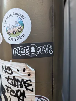 Street sticker A rectangular black sticker displaying the word 'MEGOMAR' in white bubble letters. The letter 'O' is replaced by a stylized black and white drawing of a person with long hair.