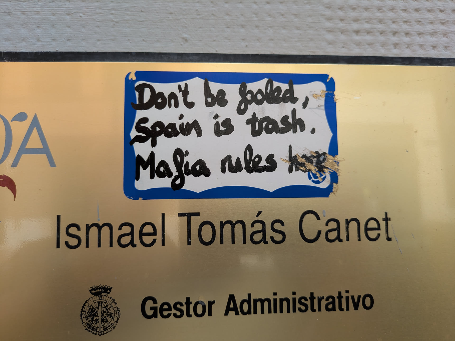 A sticker with a blue border and handwritten text in black marker. The sticker reads, "Don't be fooled, Spain is trash. Mafia rules." It's placed on a sign that reads "Ismael Tomás Canet, Gestor Administrativo."