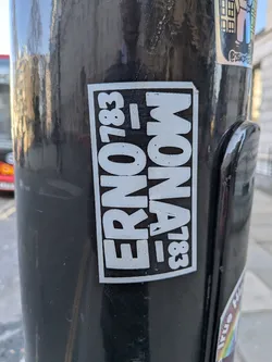 Street sticker A rectangular, black and white sticker adhered to a dark, reflective pole. The sticker features bold, sans-serif white text on a solid black background, enclosed by a thin white border. The text is arranged in two vertical columns. The left column clearly reads 'ERNO' stacked vertically, followed by '783' stacked vertically. The right column mirrors this structure but is oriented upside down relative to the left column, displaying 'MONA' stacked vertically (with the 'A' at the top and 'M' at the bottom) and '783' stacked vertically (with the '3' at the top and '7' at the bottom). The overall design is clean, graphic, and industrial in feel, with some minor wear and tear visible on the white elements, suggesting it's a physical sticker.