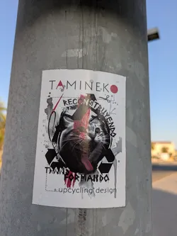 Street sticker A white sticker on a metal pole features an image of a cat and the text 'Tamineko Reconstruyendo Transformando upcycling design'. The art is done with black and red paint.
