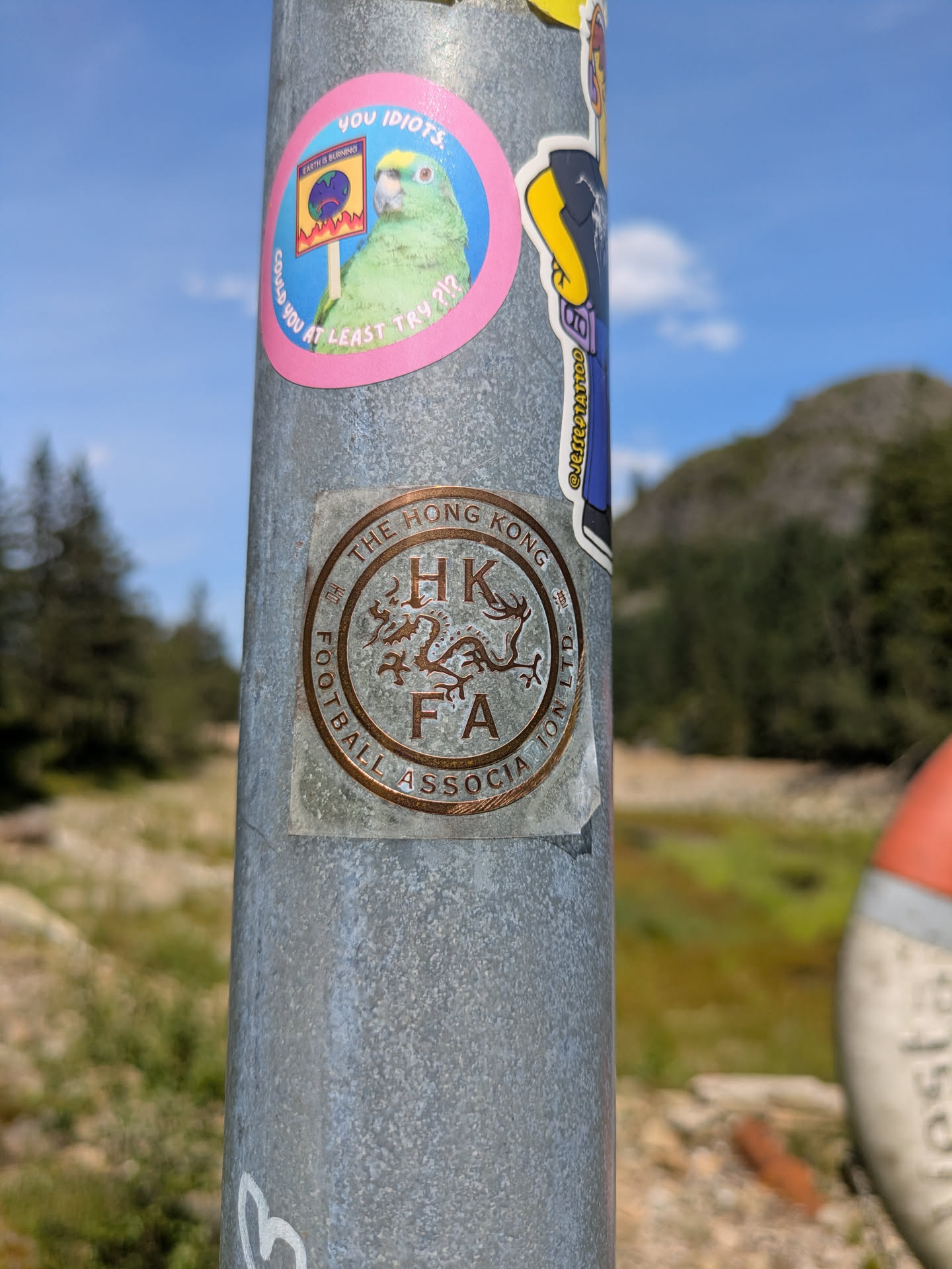 The sticker is circular with a worn, metallic bronze or copper appearance, depicting the logo of the Hong Kong Football Association. It shows initials 'HK' and 'FA' with a dragon symbol in the center, and the full name 'THE HONG KONG FOOTBALL ASSOCIATION LTD.' around the outer edge. The sticker is affixed to a textured grey metal pole and appears weathered from outdoor exposure.