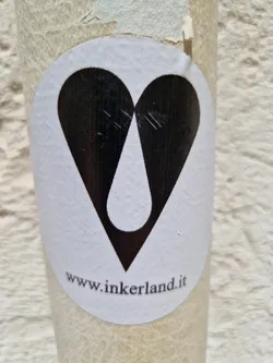 White round sticker on a beige surface with a black heart shape containing a white teardrop in its center. At the bottom, a website address is visible: www.inkerland.it