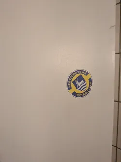 Street sticker A circular sticker featuring the logo of Hertford Town Football Club. The design consists of a blue outer ring with yellow text that reads 'HERTFORD TOWN' at the top and 'FOOTBALL CLUB' at the bottom. The inner part of the logo is a blue shield shape, which contains a white stag (deer) standing on three white wavy lines, likely representing water. The overall color scheme is blue, yellow, and white.