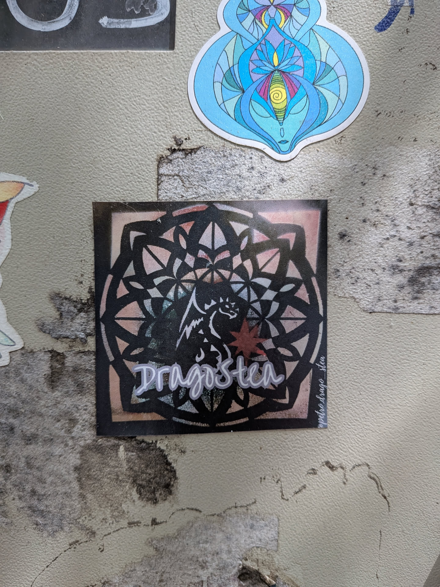 Square sticker featuring a black mandala overlay on a pink gradient. Center depicts a white dragon outline, a red star, and 'Dragostea' in white script. Includes a small social handle on the right edge.