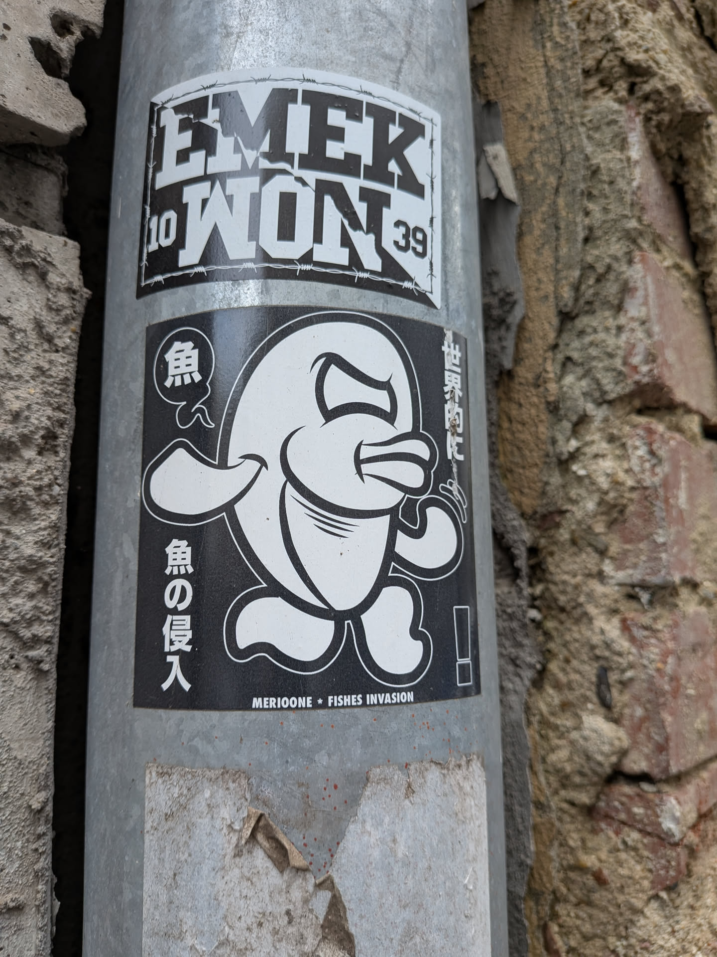 A rectangular black and white sticker depicting a stylized, angry cartoon fish with legs. It features Japanese text on the sides and 'MERIOONE * FISHES INVASION' printed along the bottom edge.