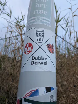 Street sticker A white square sticker divided into four quadrants by a black diagonal cross. It displays a Dubbeglas illustration at the top, the red 1. FCK logo on the left, a red devil head on the right, and the text 'Dubbe Deiwel' at the bottom.