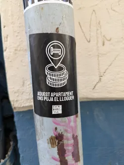 The sticker features a bed in a map pointer above two stacks of coins, and the text "AQUEST APARTAMENT ENS PUJA EL LLOGUER", which translates to "This apartment is raising our rent". The sticker is from Entre Barris VLC.