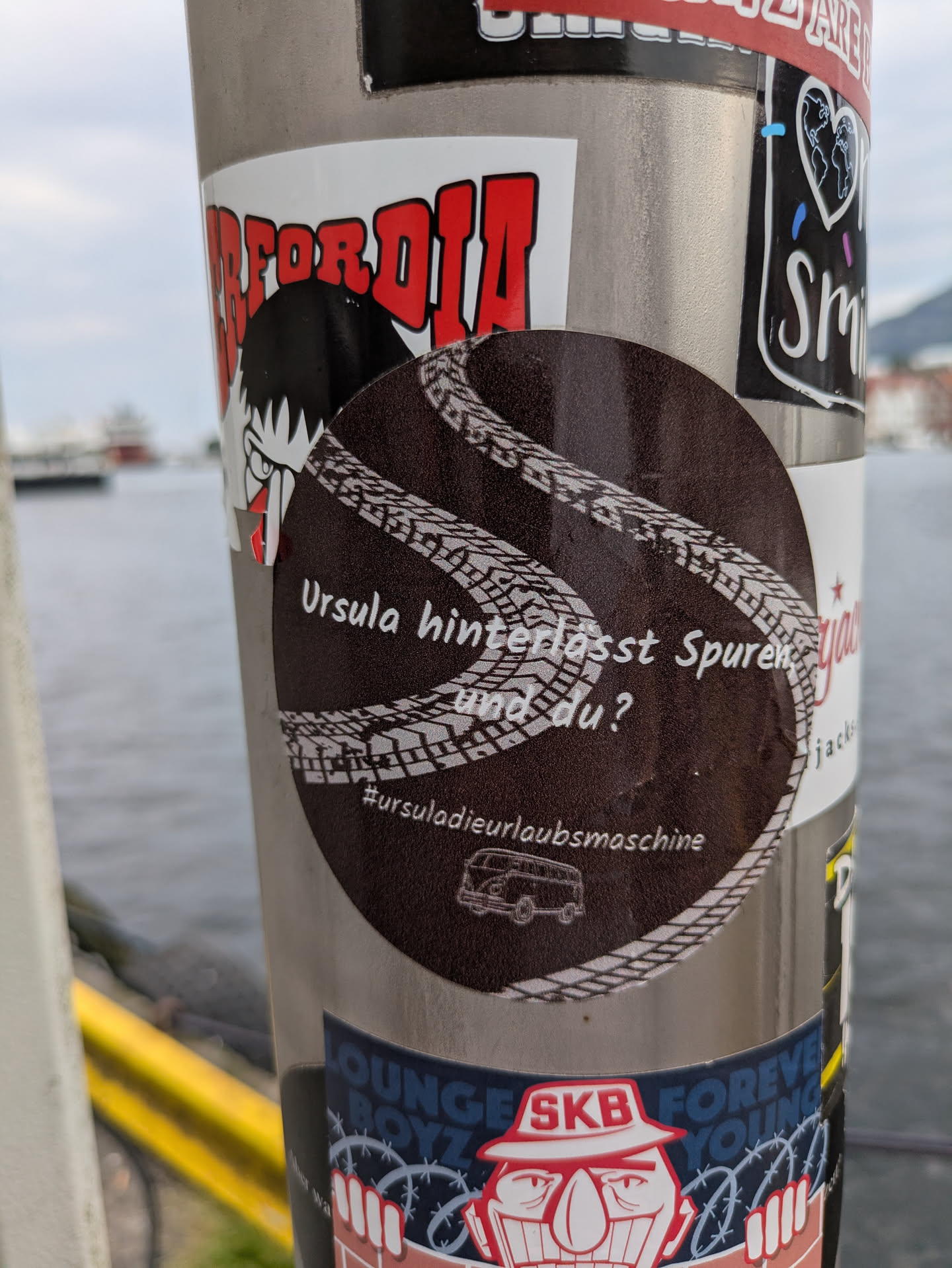 The central sticker is circular and dark brown. It features two prominent white tire track patterns that curve across its surface. In the center, white text in German reads 'Ursula hinterlässt Spuren und du?' (Ursula leaves traces, and you?). Below this text, there is a hashtag '#ursuladieurlaubsmaschine' and a small, white outline illustration of a vintage camper van, reminiscent of a VW Bus. The sticker is placed on a metal pole, with other stickers partially visible around it.