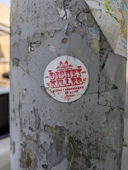 Street sticker The sticker is white and circular with all text and decorative elements in red. At the top, in smaller text, it reads "immaturus". Below this, the dominant text in a distressed, bold font says "DISNEY KILLER". Further down, in smaller text, it states "spilles i storelogen 30 april", suggesting an event or performance. The sticker has ornate decorative borders above and below the main text. It is placed on a heavily textured, peeling grey pole with visible signs of age and wear.