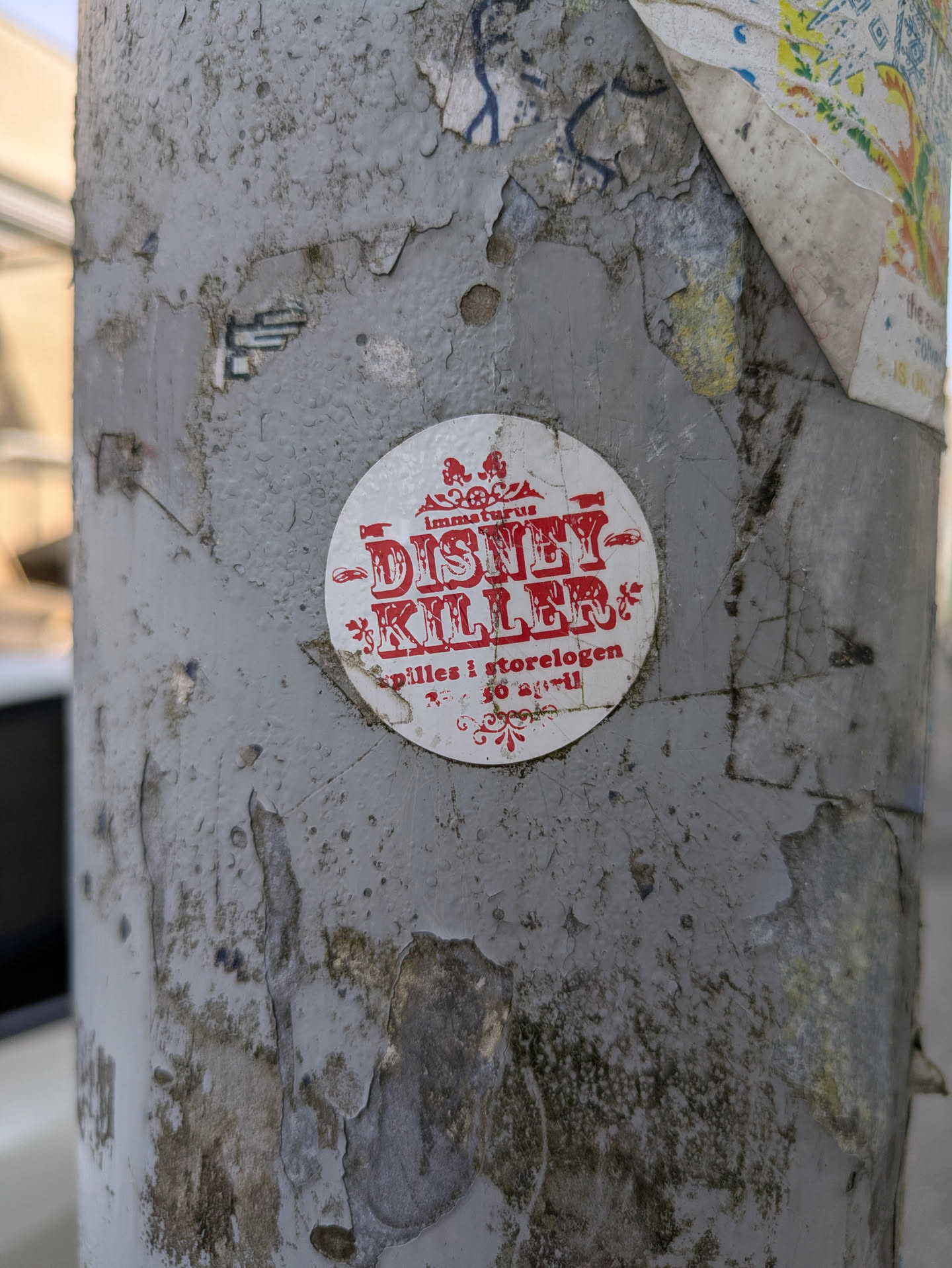 The sticker is white and circular with all text and decorative elements in red. At the top, in smaller text, it reads "immaturus". Below this, the dominant text in a distressed, bold font says "DISNEY KILLER". Further down, in smaller text, it states "spilles i storelogen 30 april", suggesting an event or performance. The sticker has ornate decorative borders above and below the main text. It is placed on a heavily textured, peeling grey pole with visible signs of age and wear.
