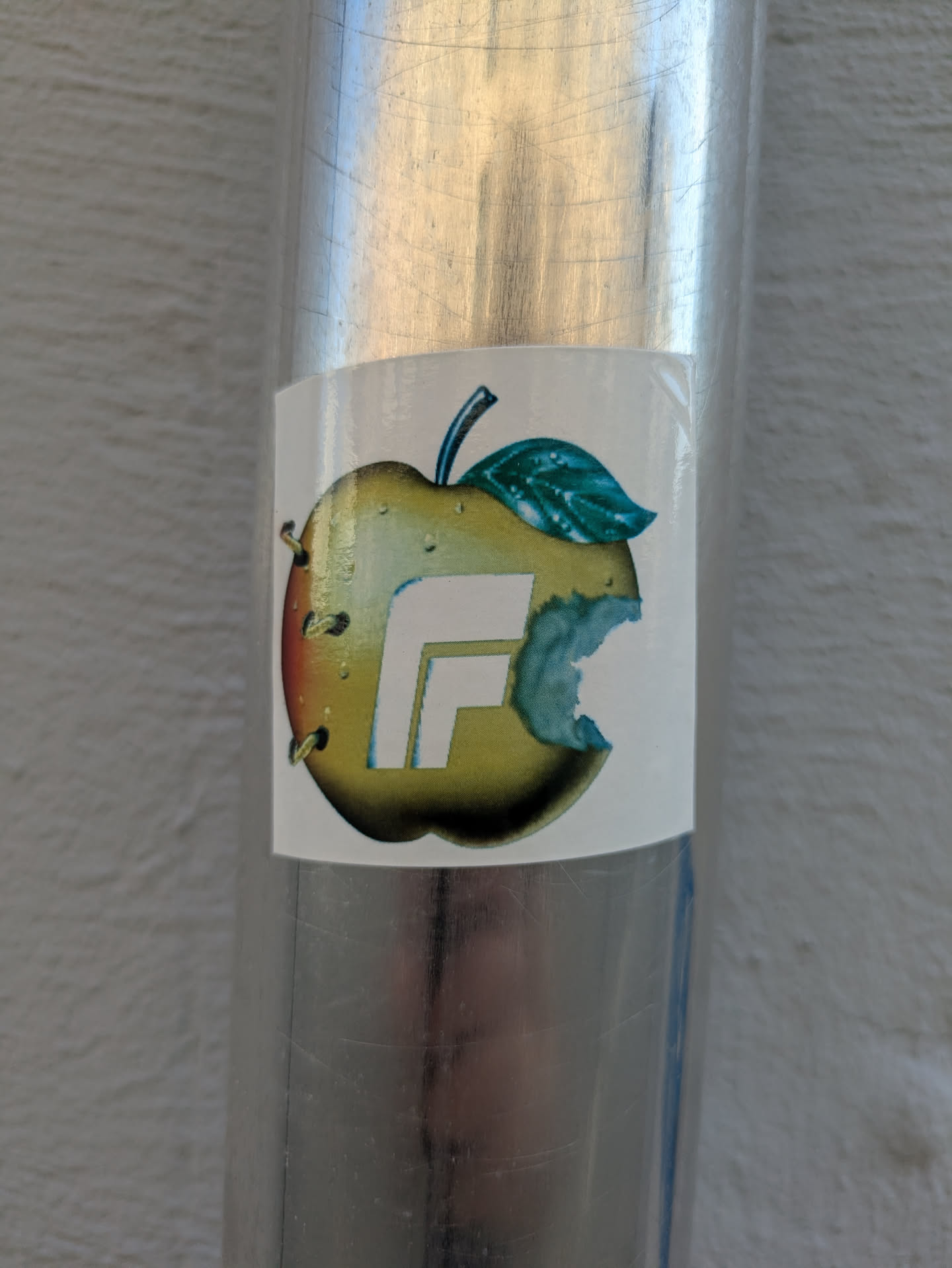 The sticker depicts a stylized apple, primarily yellow-green with a subtle reddish blush on its left side. A distinct bite mark is visible on the right side of the apple. A large, bold, white letter 'F' is integrated into the center of the apple design. A green leaf and a dark stem protrude from the top. On the left side, there are a few small, dark, circular elements that could represent seeds or small piercings. The sticker is affixed to a shiny, scratched metallic cylindrical surface.
