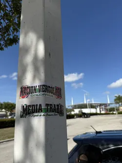 Street sticker Two rectangular stickers are affixed to a light-colored concrete pole. The top sticker features the bold, black text 'NACION VERDOLAGA' with a green outline, and 'COLOMBIA' in smaller black letters below it. The background of this sticker is a collage of colors including red, yellow, blue, and green, possibly incorporating elements of the Colombian flag and football club motifs. The bottom sticker displays the prominent black text 'MEDIA TRABA' with a green outline, and below it, 'El Legado de las Primera Linea' in a smaller, stylized script. Its background is a mix of green, black, and white patterns, with some leaf-like designs on the sides. Both stickers show slight wear and tear, consistent with outdoor placement.