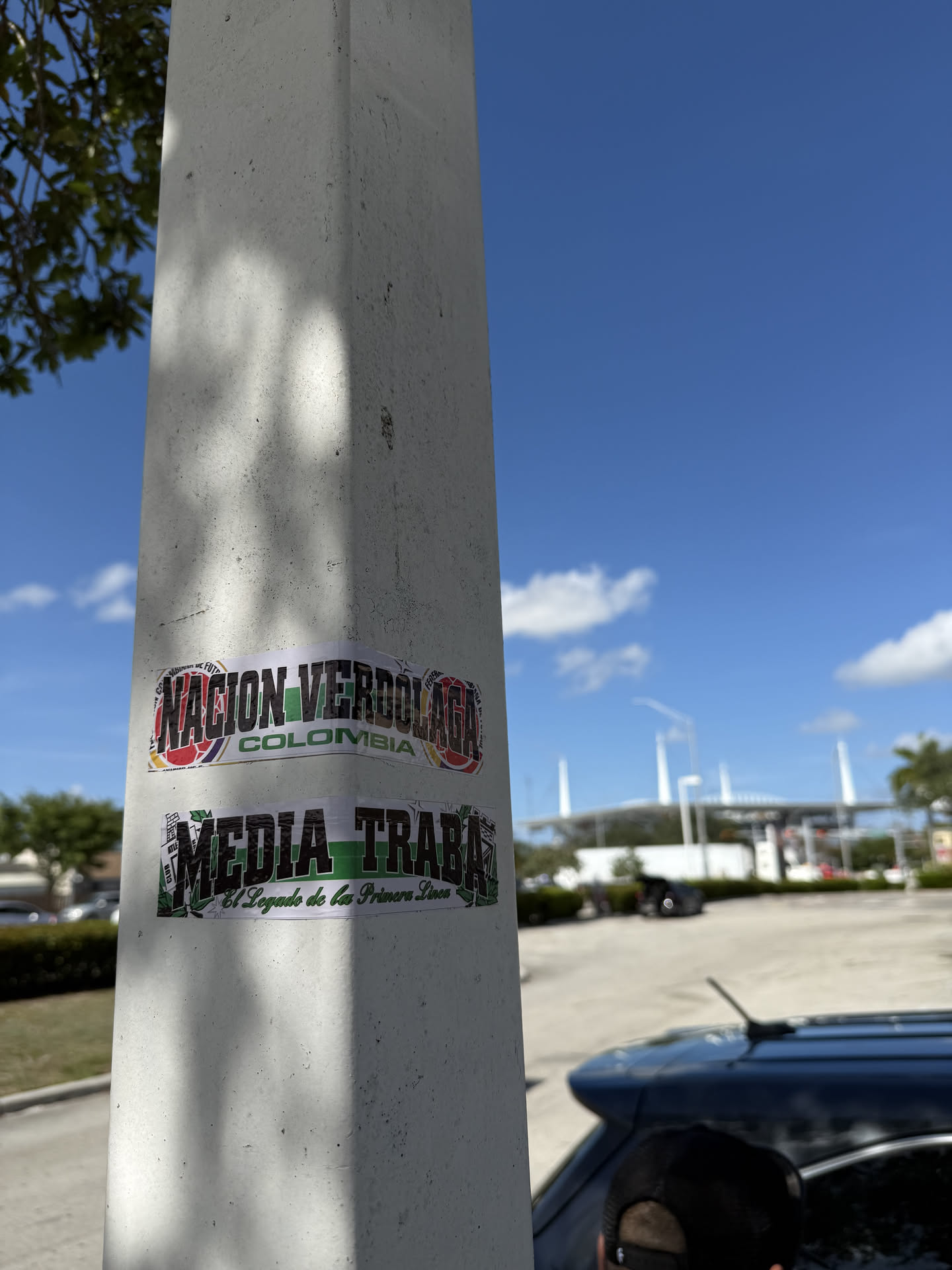 Two rectangular stickers are affixed to a light-colored concrete pole. The top sticker features the bold, black text 'NACION VERDOLAGA' with a green outline, and 'COLOMBIA' in smaller black letters below it. The background of this sticker is a collage of colors including red, yellow, blue, and green, possibly incorporating elements of the Colombian flag and football club motifs. The bottom sticker displays the prominent black text 'MEDIA TRABA' with a green outline, and below it, 'El Legado de las Primera Linea' in a smaller, stylized script. Its background is a mix of green, black, and white patterns, with some leaf-like designs on the sides. Both stickers show slight wear and tear, consistent with outdoor placement.