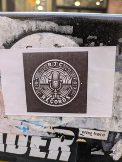 This sticker shows the logo for RJC Records, featuring a vintage-style microphone inside a circle. The seal is decorated with stars and geometric patterns and has a retro feel. The background is dark brown.