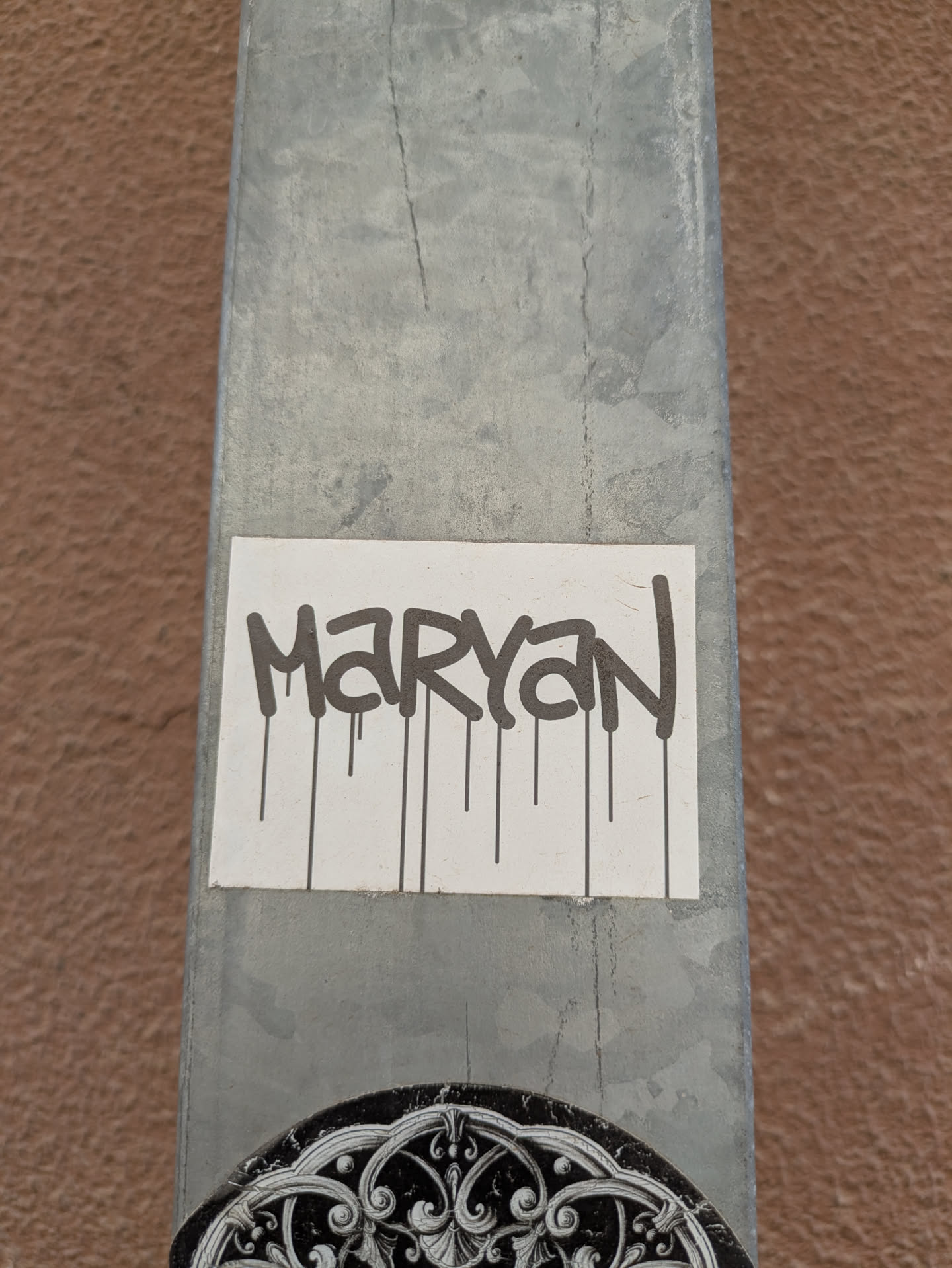 A sticker is adhered to a gray metal pole. The sticker has the name "MARYAN" written in black, dripping style letters on a white background. Another sticker featuring a decorative black and white design is partially visible below it.