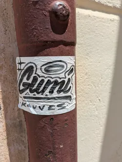A white sticker with black, stylized graffiti lettering that reads 'Gumi' and underneath, '‘Kauves’'. The sticker is attached to a red metal pipe.