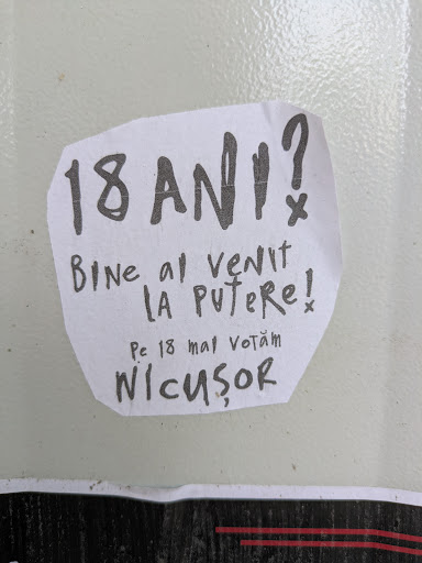 The image shows a hand-written sticker with a political message in Romanian, featuring the name 'Nicușor'. The sticker appears to be promoting a candidate, as it mentions voting on May 18th. The overall aesthetic suggests a grassroots or DIY campaign effort.