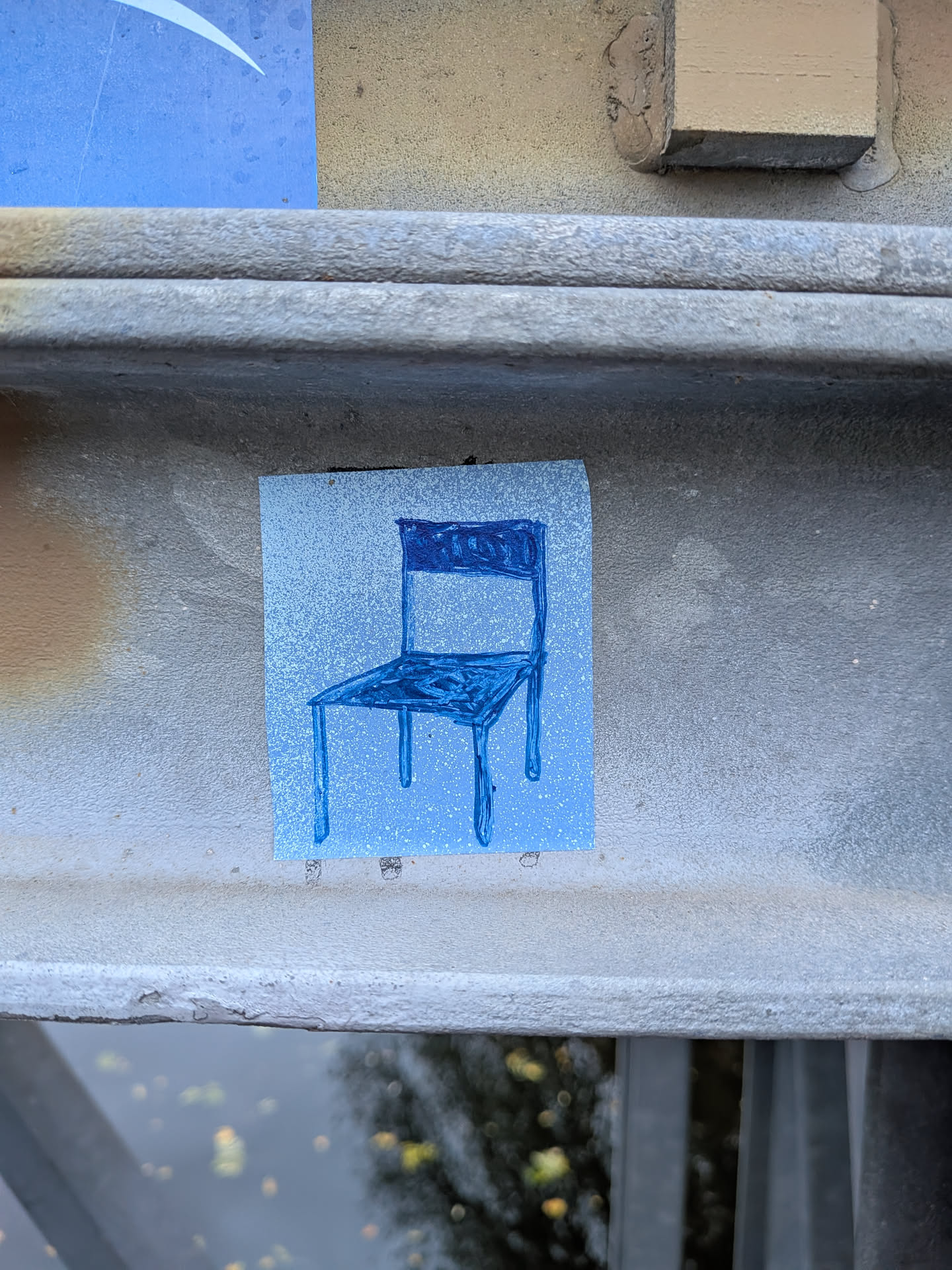 The sticker is square-ish and features a simple, hand-drawn illustration of a chair. The chair is depicted in a dark blue outline and filled with a lighter blue, with some darker blue accents on the backrest and seat. The background of the sticker is a light blue with a subtle speckled texture, resembling spray paint. The overall style is minimalist and somewhat childlike.