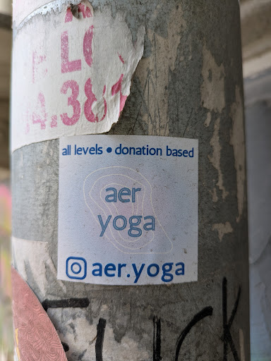 A square sticker for AER Yoga is adhered to a pole. The sticker reads "all levels • donation based aer yoga @aer.yoga" followed by the instagram logo.