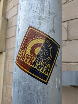 Street sticker The sticker is square-shaped with rounded corners and is affixed to a metal pole. It features a design split diagonally. The upper left portion is a deep brown with a yellow concentric circle and a stylized 'C' shape within it. The lower right portion is black with yellow lines forming a circular pattern that resembles a stylized eye or a target, with radiating lines. The text 'ÖSTRSTA' is prominently displayed in a yellow, stylized font across the center of the sticker, overlaying both color sections. The sticker shows signs of wear, with some creasing and small white specks, suggesting it has been exposed to the elements.