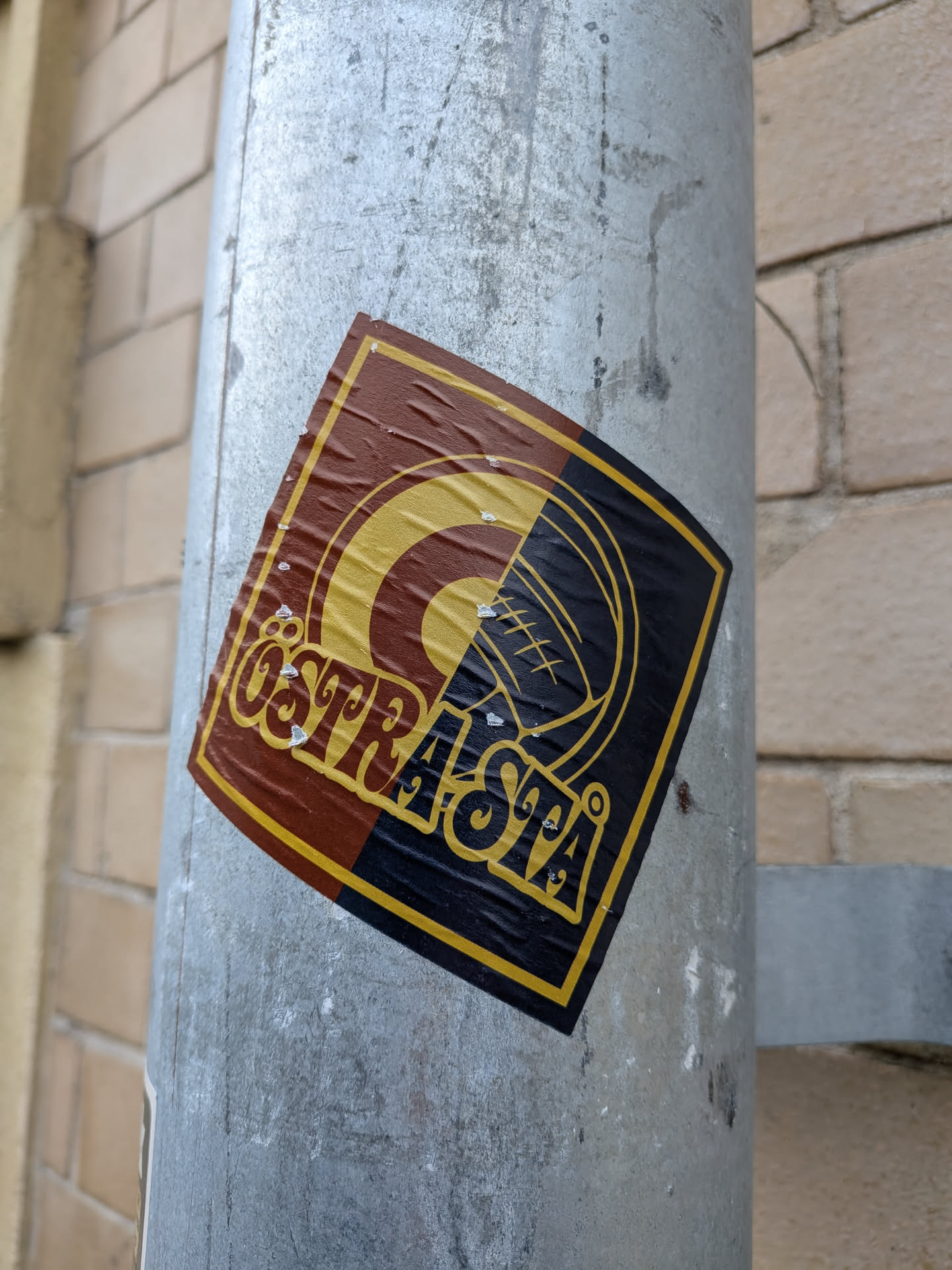 The sticker is square-shaped with rounded corners and is affixed to a metal pole. It features a design split diagonally. The upper left portion is a deep brown with a yellow concentric circle and a stylized 'C' shape within it. The lower right portion is black with yellow lines forming a circular pattern that resembles a stylized eye or a target, with radiating lines. The text 'ÖSTRSTA' is prominently displayed in a yellow, stylized font across the center of the sticker, overlaying both color sections. The sticker shows signs of wear, with some creasing and small white specks, suggesting it has been exposed to the elements.