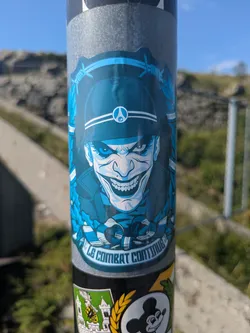 Street sticker The sticker showcases a stylized, blue-toned illustration of a character with a menacing grin, resembling the Joker, wearing a dark blue military-style helmet. The helmet features a white circular emblem with a symbol inside, possibly a stylized 'G' or a peace-like sign. The character has sharp teeth, a nose piercing, and piercing eyes. Barbed wire frames the top part of the sticker, and leafy patterns in lighter blue tones are visible on the sides. At the bottom, a curved banner contains the text "LE COMBAT CONTINUE" in white capital letters. The overall design conveys a sense of grim determination and a somewhat unsettling aesthetic.