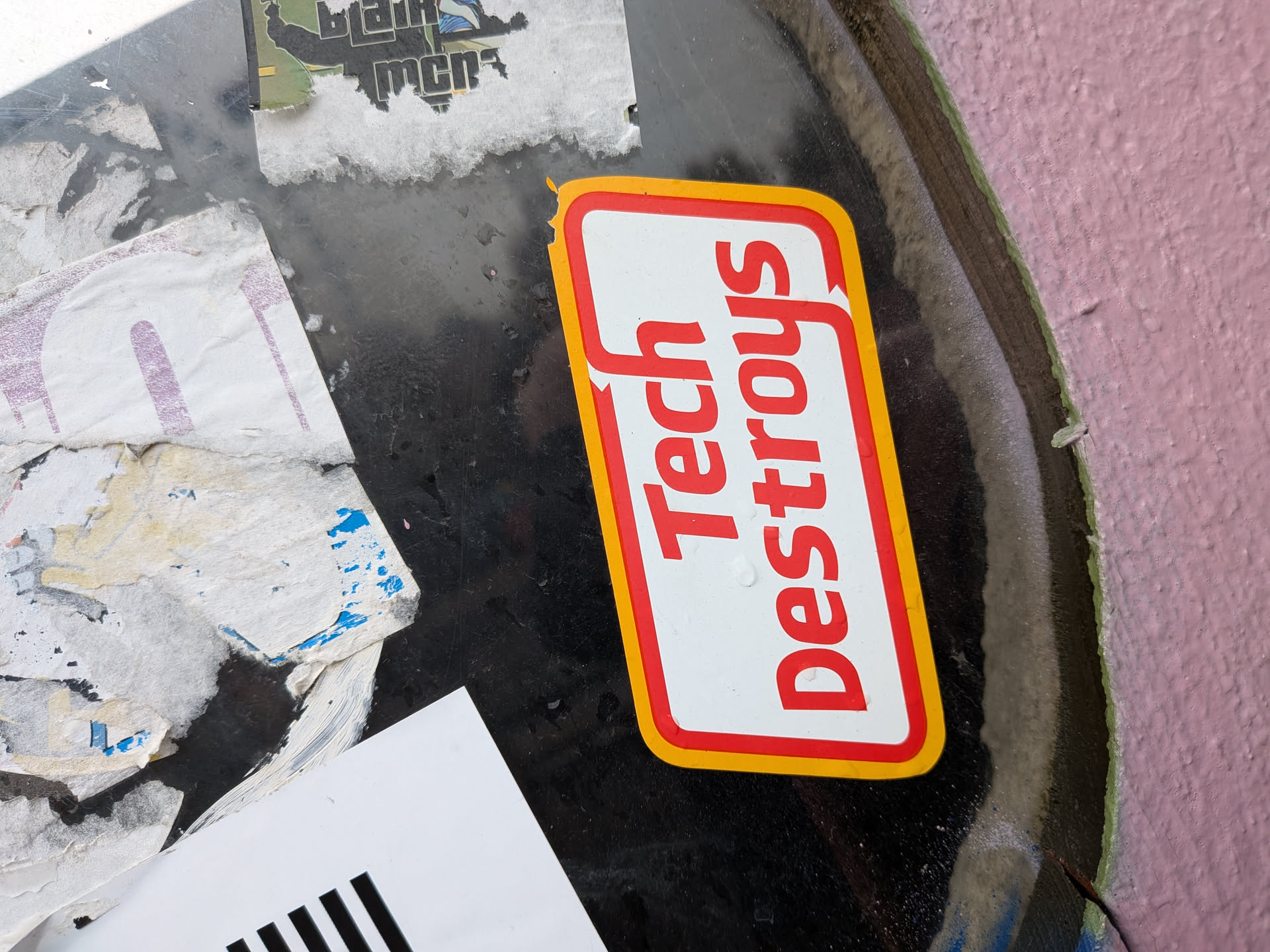 The sticker is rectangular with rounded corners and features a prominent red border and a thinner yellow border around it. The background of the sticker is white. Bold, red, sans-serif text reads 'Tech Destroys' vertically. The text is slightly angled, and the letters are large and impactful. There are a few small water droplets or smudges on the white surface of the sticker. The sticker appears to be slightly peeling at the top left corner.