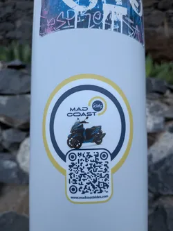Street sticker Circular white sticker with a yellow and dark blue border. Features a blue three-wheeled scooter in the center, the text 'MAD COAST Rides', and a QR code at the bottom with the website 'www.madcoastrides.com'.