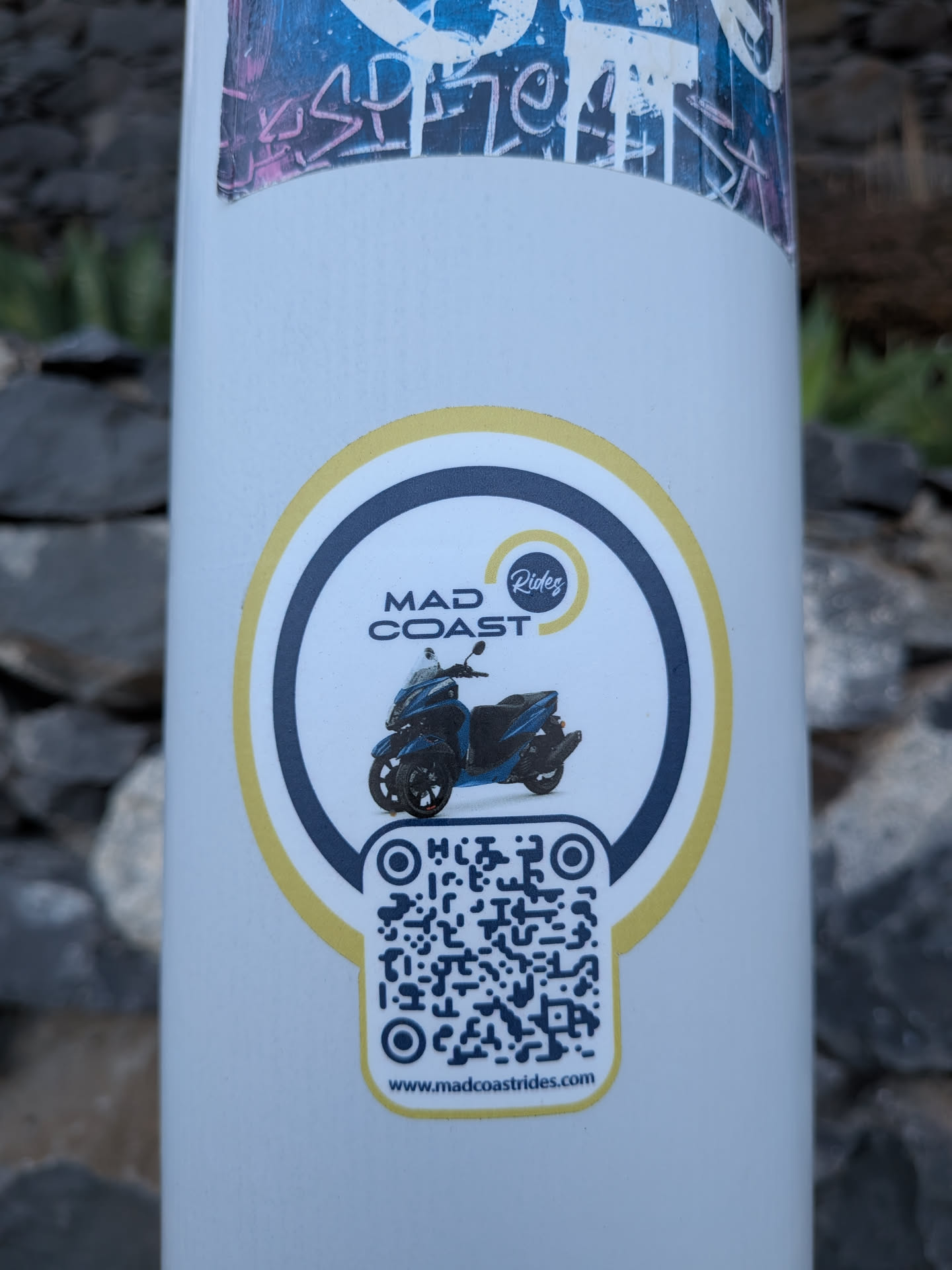 Circular white sticker with a yellow and dark blue border. Features a blue three-wheeled scooter in the center, the text 'MAD COAST Rides', and a QR code at the bottom with the website 'www.madcoastrides.com'.